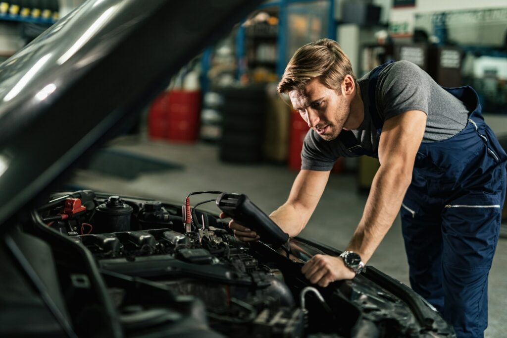 Young mechanic with diagnostic tool analyzing car engine problem in auto repair shop.